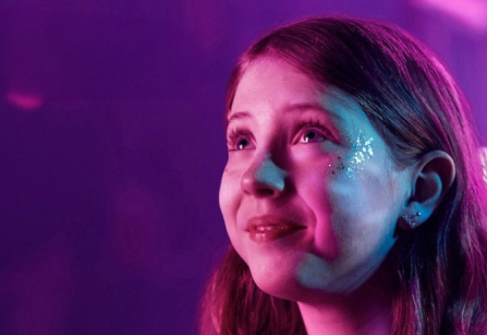 A girl with glitter on her face, possibly at a concert, looks up with a smile. The image has a purple and pink color scheme