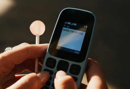 A person holds a simple, blue, non-smartphone in their hands. They are in the process of typing a message that says "Hello, I'm so sad." on the small screen. In their left hand, they are also holding a pink lollipop.