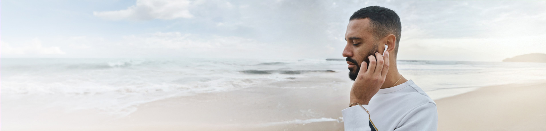 A man, seen in profile, is standing on a beach wearing a white long-sleeved shirt and an earbud in his right ear, and he is touching the side of his face. The background is a calm beach with waves gently washing the shore under a soft, overcast sky.