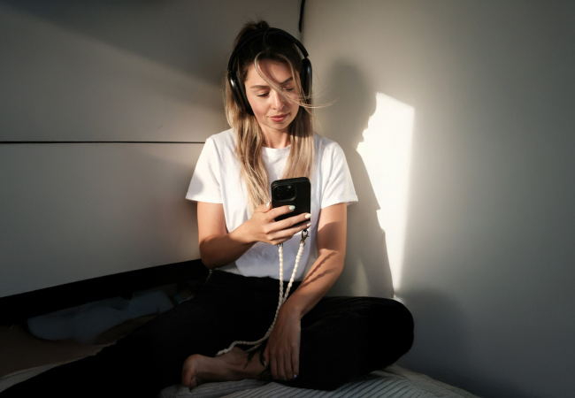 A young woman is sitting on a bed in a sunlit room, with her legs crossed. She is wearing headphones and holding a smartphone and looking down at the screen. A white beaded strap is attached to her phone.