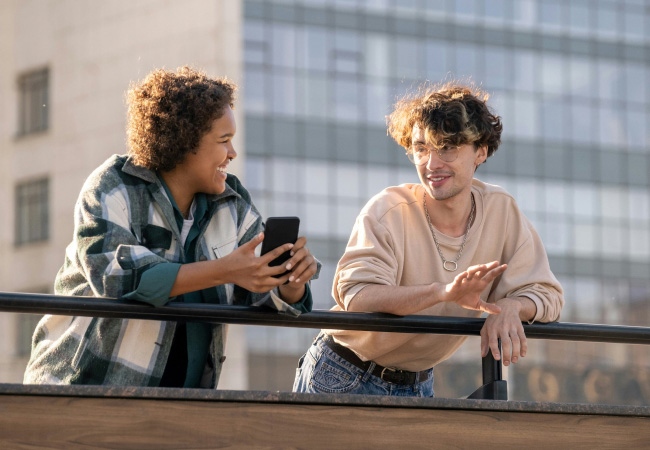 Two friends are leaning against a black railing on a sunny day. One person is looking down at their phone and laughing. The other is looking at them and talking, with a slight smile on their face.